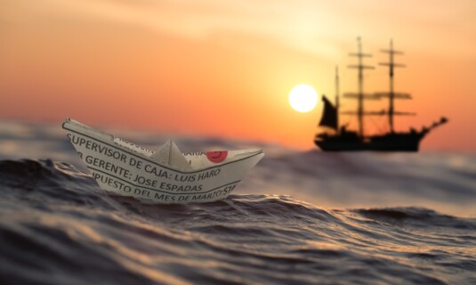 Papierschiff auf Wellen, im Hintergrund ein Segelschiff im Sonnenuntergang